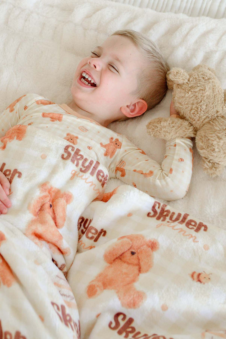 Smiling boy in Caden Lane honey bear print pajamas laying on a bed, holding a plush teddy bear with a personalized Caden Lane blanket printed with 'Skyler Quinn'