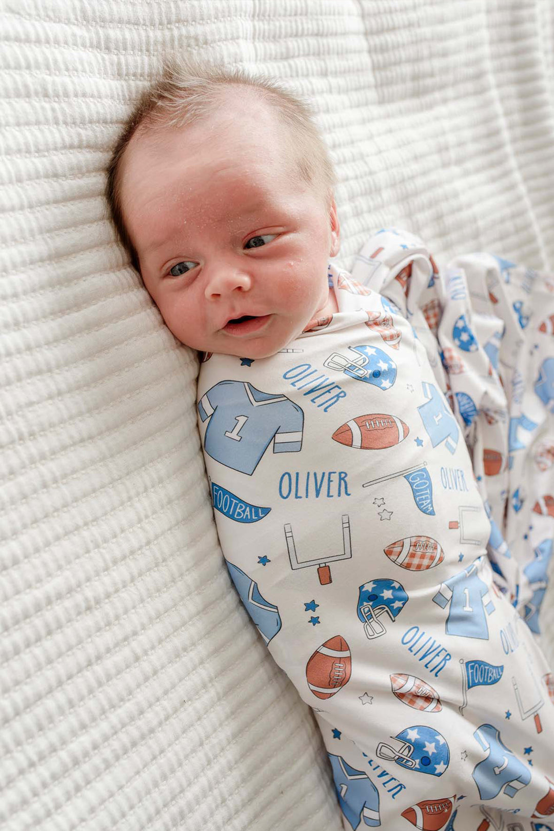 close up of a baby wrapped in a Caden Lane huddles and cuddles blue football swaddle. 