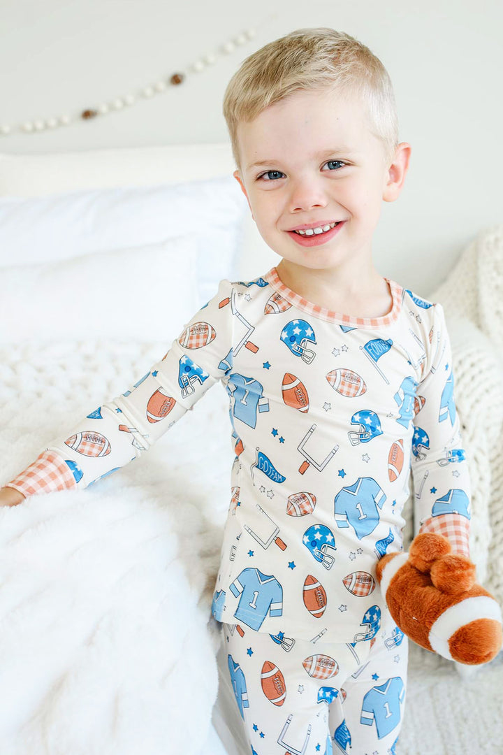 Little boy standing next to a bed and wearing the Caden Lane blue huddles and cuddles two piece pajama set while holding a football stuffy. 