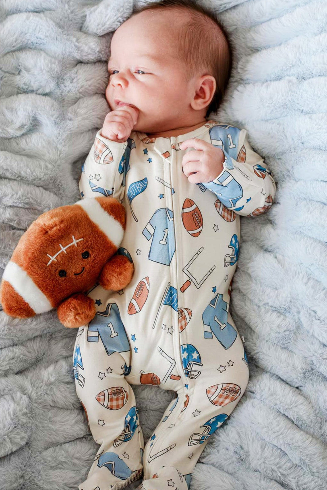 A baby lying on a textured light blue blanket, wearing a cream-colored footed onesie with a blue and brown football pattern, and holding a brown plush football toy.