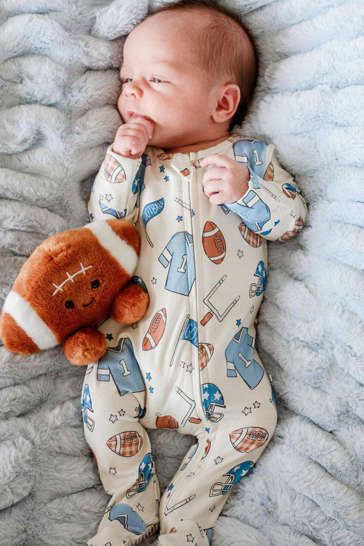 A baby lying on a textured light blue blanket, wearing a cream-colored footed onesie with a blue and brown football pattern, and holding a brown plush football toy.