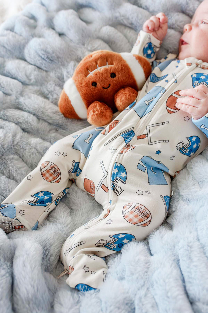 Newborn baby laying on a blue soft blanket while wearing the Caden Lane huddles & cuddles blue zip romper. The print features football prints! 