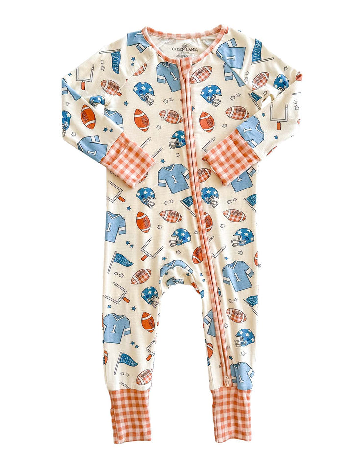A baby long-sleeve footless one-piece sleeper with a sports-themed print of blue helmets, orange footballs, and white tennis rackets on a cream background. It features orange and white gingham cuffs and a matching orange zipper trim.
