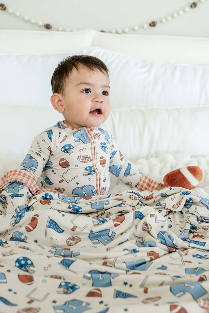 Baby sitting and holding a stuffed football on a white fuzzy blanket, wearing a Caden Lane 'Huddles and Cuddles' blue football-print outfit with orange gingham trim, with matching bamboo blanket laying on his legs