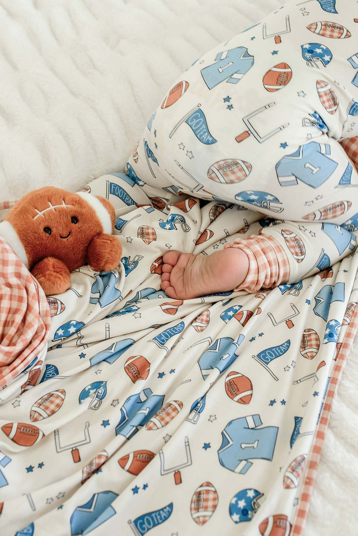 Close-up of a baby's bare foot peeking out from a Caden Lane 'Huddles and Cuddles' blue football-print blanket, next to a plush football stuffed toy on a white fuzzy blanket