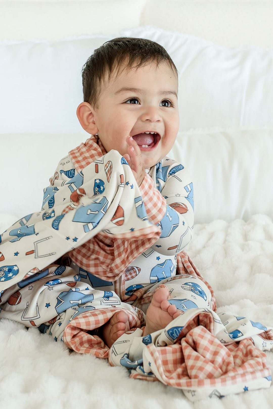 Laughing baby sitting on a white fuzzy blanket, wearing a Caden Lane 'Huddles and Cuddles' blue football-print outfit with orange gingham trim, holding a matching bamboo blanket