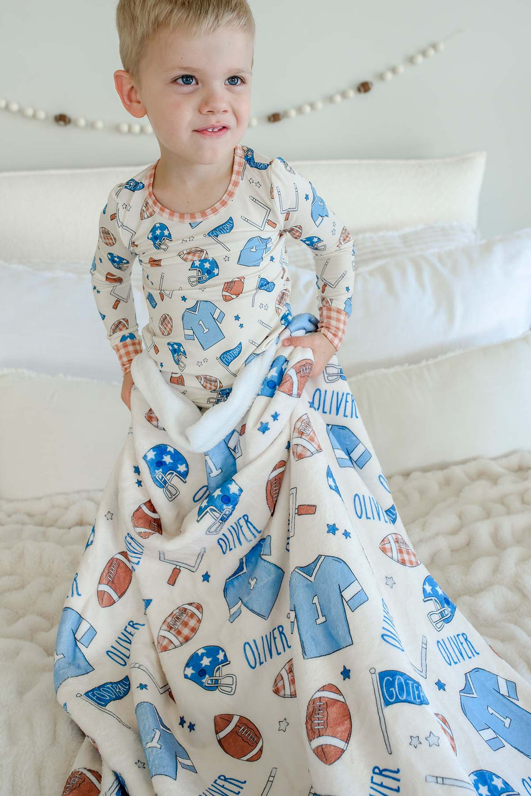 Smiling young boy standing on a cream faux-fur blanket, wearing football-print pajamas, snuggled with a personalized Caden Lane 'Huddles and Cuddles' blue minky blanket featuring football helmets, jerseys, and the name Oliver