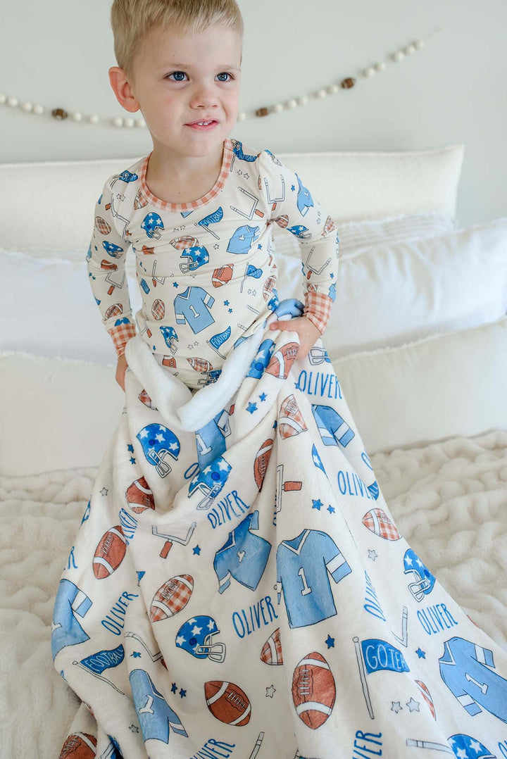 Smiling young boy standing on a cream faux-fur blanket, wearing football-print pajamas, snuggled with a personalized Caden Lane 'Huddles and Cuddles' blue minky blanket featuring football helmets, jerseys, and the name Oliver