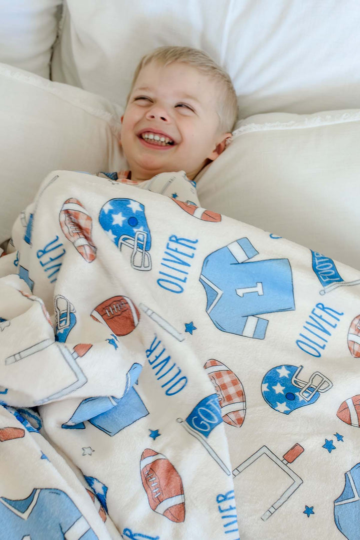 Smiling young boy lying on a cream bed,  snuggled with a personalized Caden Lane 'Huddles and Cuddles' blue minky blanket featuring football helmets, jerseys, and the name Oliver