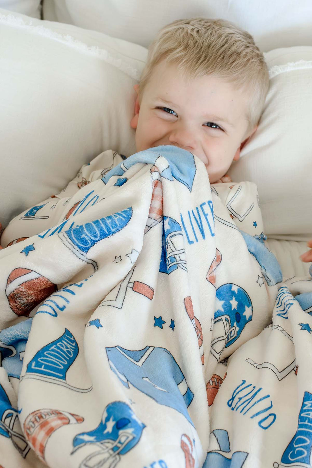 Boy lying on a cream bed,  snuggled with a personalized Caden Lane 'Huddles and Cuddles' blue minky blanket featuring football helmets, jerseys, and the name Oliver
