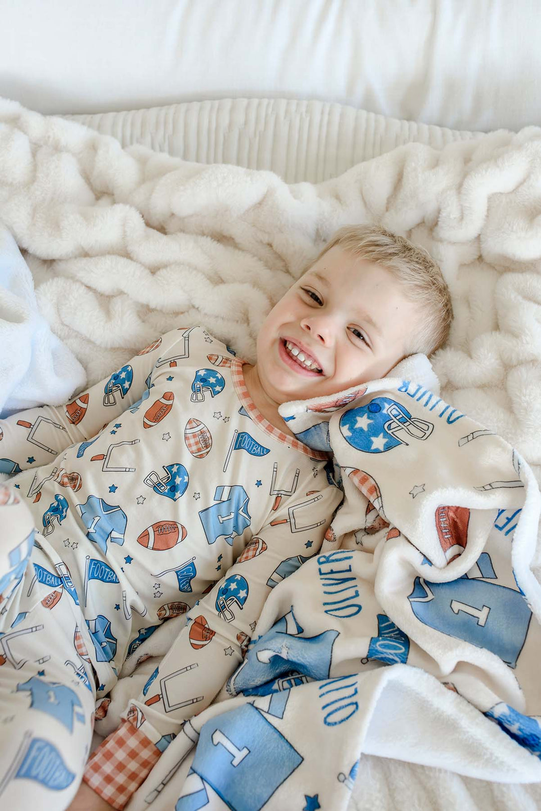 Smiling young boy lying on a cream faux-fur blanket, wearing football-print pajamas, snuggled with a personalized Caden Lane 'Huddles and Cuddles' blue minky blanket featuring football helmets, jerseys, and the name Oliver