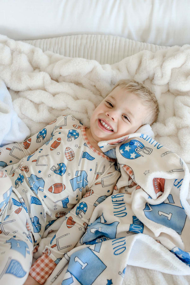 Smiling young boy lying on a cream faux-fur blanket, wearing football-print pajamas, snuggled with a personalized Caden Lane 'Huddles and Cuddles' blue minky blanket featuring football helmets, jerseys, and the name Oliver