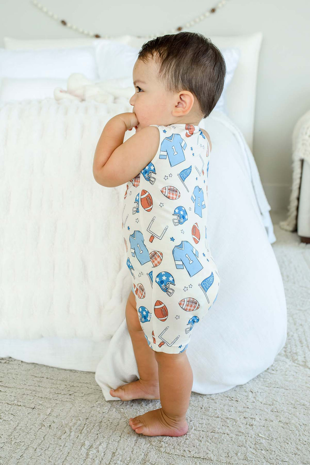 Baby standing barefoot on a rug, wearing a Caden Lane 'Huddles and Cuddles' blue football-print sleeveless romper, leaning on a bed