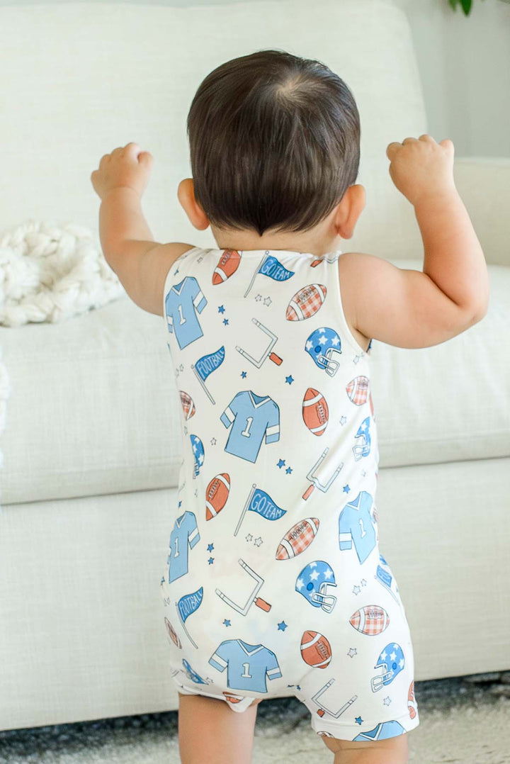 Baby turned around standing wearing a Caden Lane 'Huddles and Cuddles' blue football-print sleeveless romper