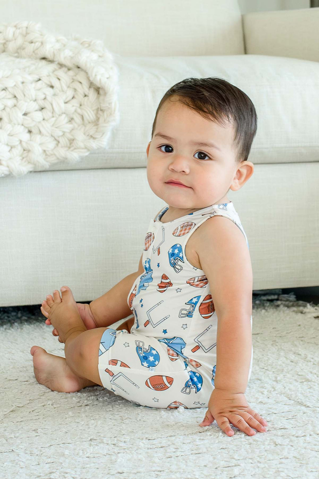 Baby sitting on a rug, wearing a Caden Lane 'Huddles and Cuddles' blue football-print sleeveless romper