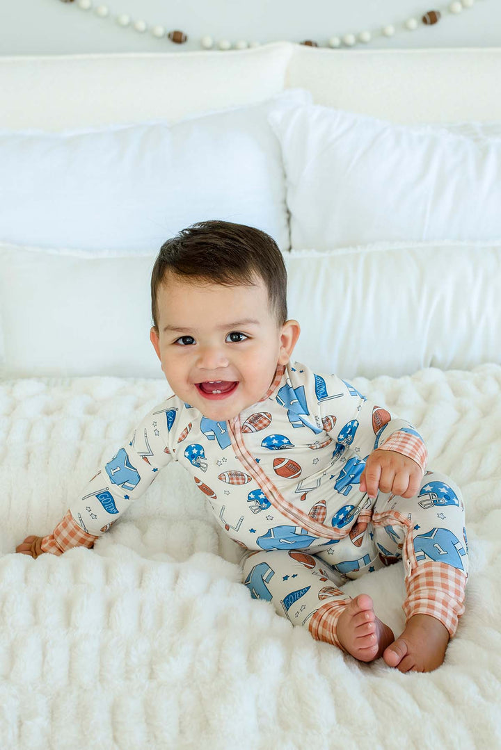 A baby sitting on a white textured blanket in front of white pillows, wearing a long-sleeve one-piece pajama with a blue whale print on a cream background and orange plaid cuffs.