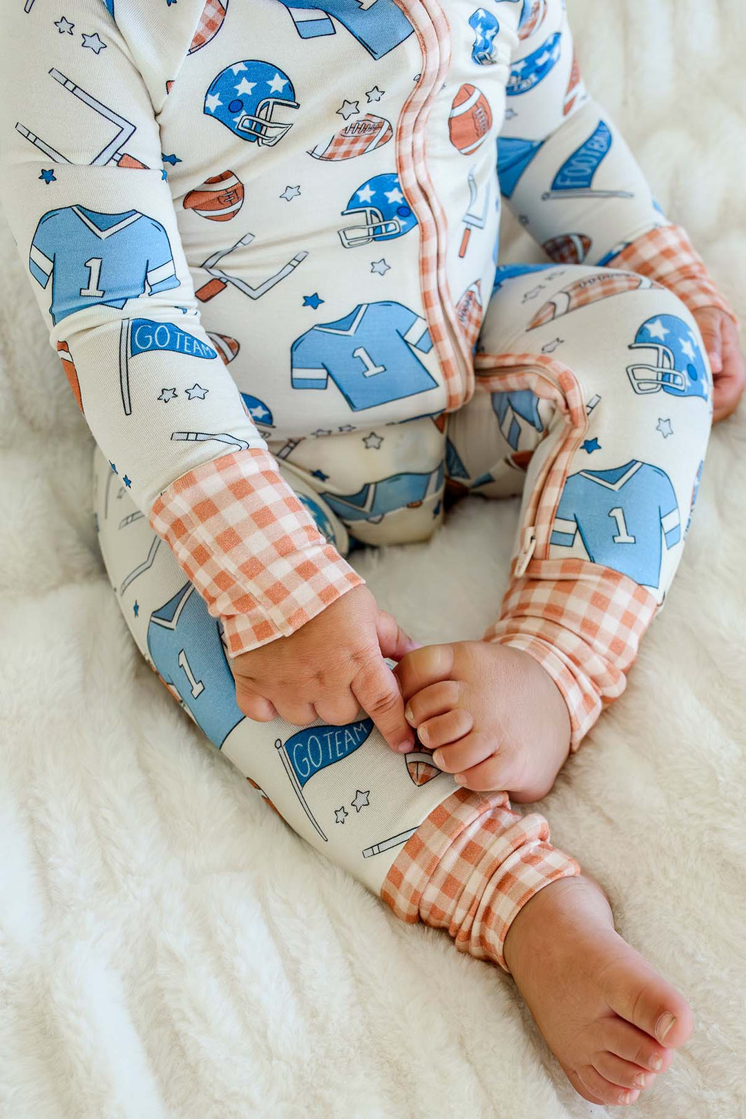 A baby wearing a long-sleeve zip-up pajama top and matching pants with a sports-themed print, sitting on a soft white blanket. The pajamas feature blue and orange football jersey graphics on a white background, with orange and white gingham cuffs.