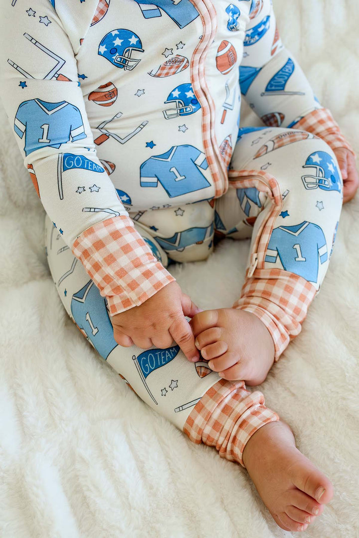 A baby wearing a long-sleeve zip-up pajama top and matching pants with a sports-themed print, sitting on a soft white blanket. The pajamas feature blue and orange football jersey graphics on a white background, with orange and white gingham cuffs.