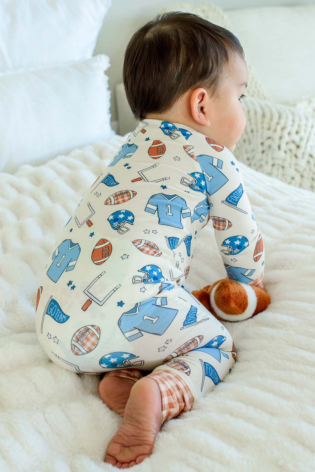 A baby sitting on a white blanket, wearing a long-sleeve footed pajama with a sports-themed print of blue jerseys, footballs, and helmets on an off-white background.