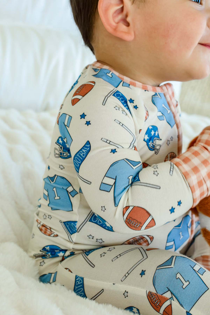 A close-up side view of a baby wearing a long-sleeve footless pajama top and matching pants with a sports-themed print on a white textured blanket.