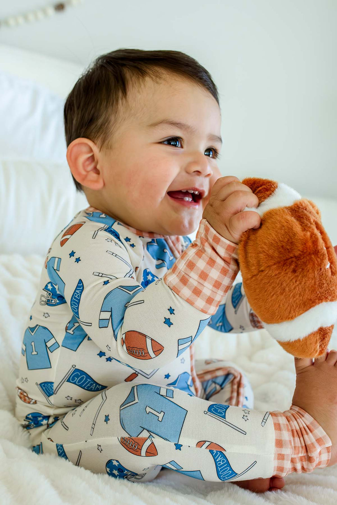 A baby sitting on a white blanket, wearing a long-sleeve pajama shirt and pants set with a sports-themed print, holding a plush football toy.
