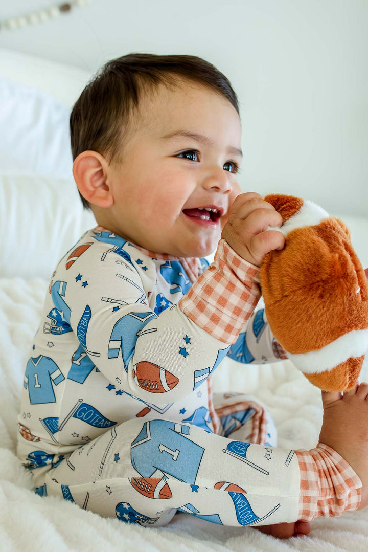 A baby sitting on a white blanket, wearing a long-sleeve pajama shirt and pants set with a sports-themed print, holding a plush football toy.