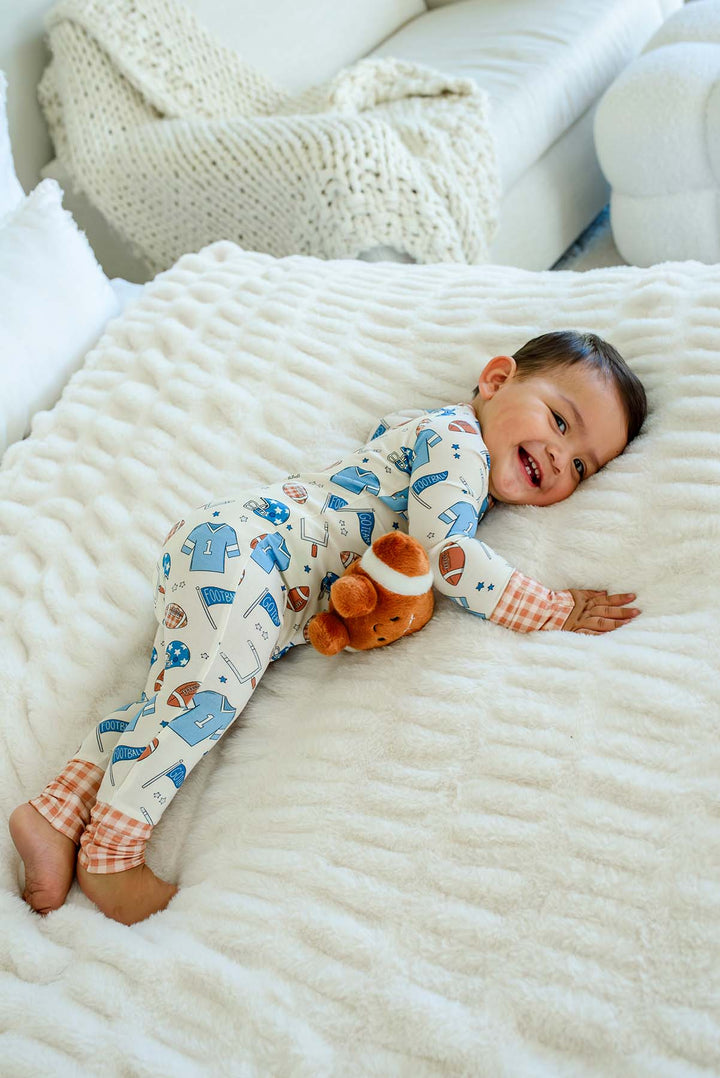 A baby lying on a textured white blanket in a cozy room, wearing a cream-colored long-sleeve pajama set with blue house prints and orange plaid cuffs, while holding an orange plush toy.