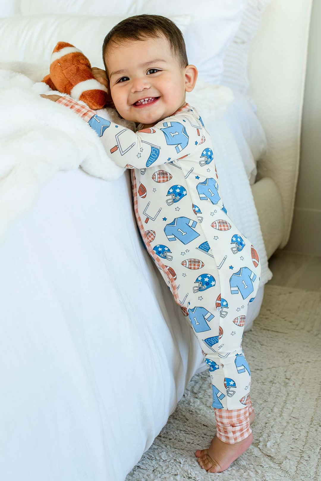 A baby standing on a carpeted floor next to a bed, wearing a cream-colored footed pajama with a blue and orange space-themed print. The baby is holding a plush toy.