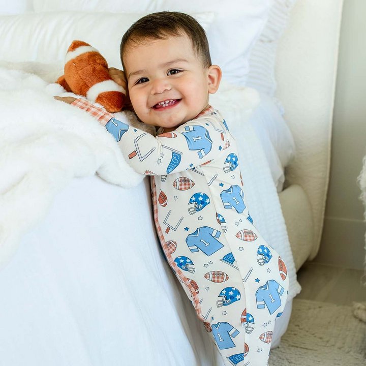 Smiling toddler leaning on a bed beside a plush toy, wearing a Caden Lane white bamboo convertible zip romper in a Huddles & Cuddles Blue print featuring footballs and jerseys.