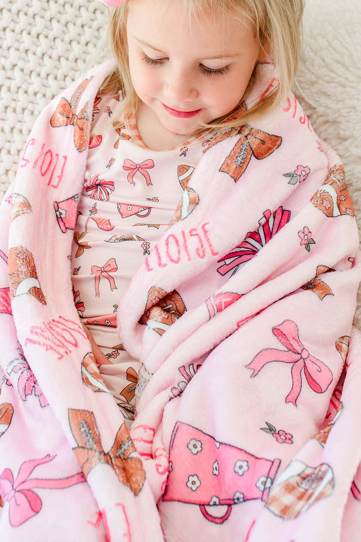 Girl wrapped in a personalized Caden Lane 'Huddles and Cuddles' pink football and cheerleading print blanket with the name Eloise, sitting