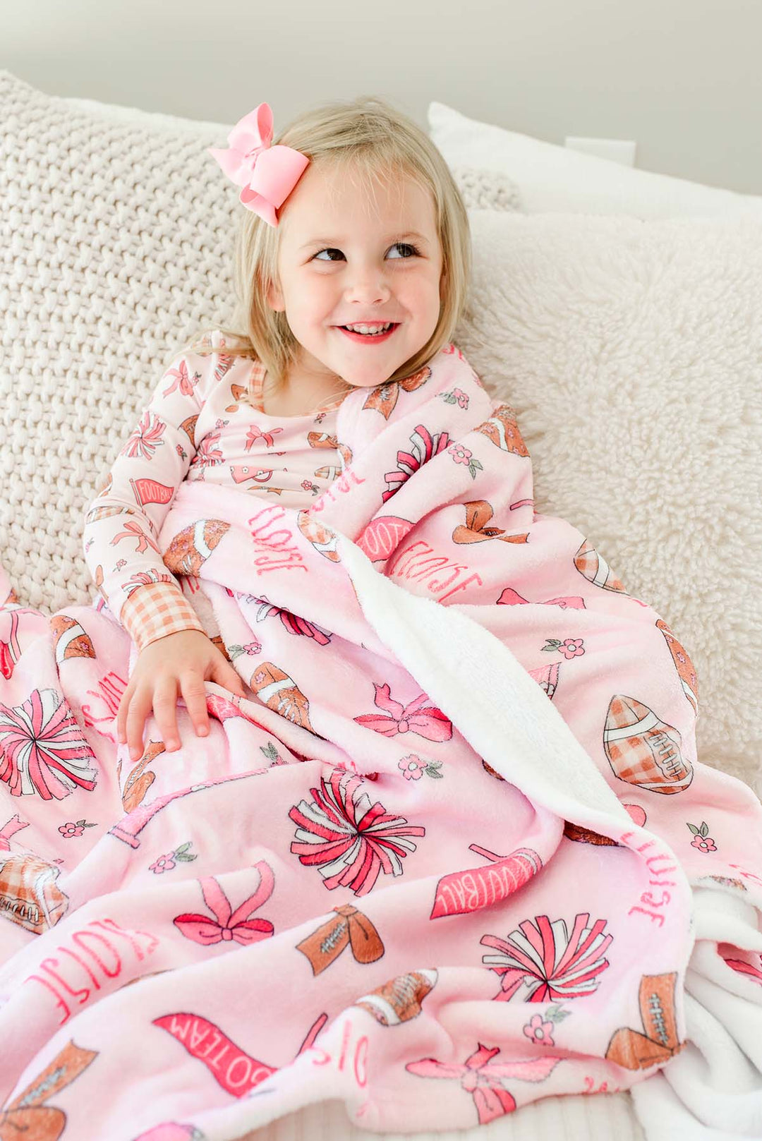 Smiling young girl with a pink hair bow, wrapped in a personalized Caden Lane 'Huddles and Cuddles' pink football and cheerleading print blanket with the name Eloise, sitting against a knit pillow