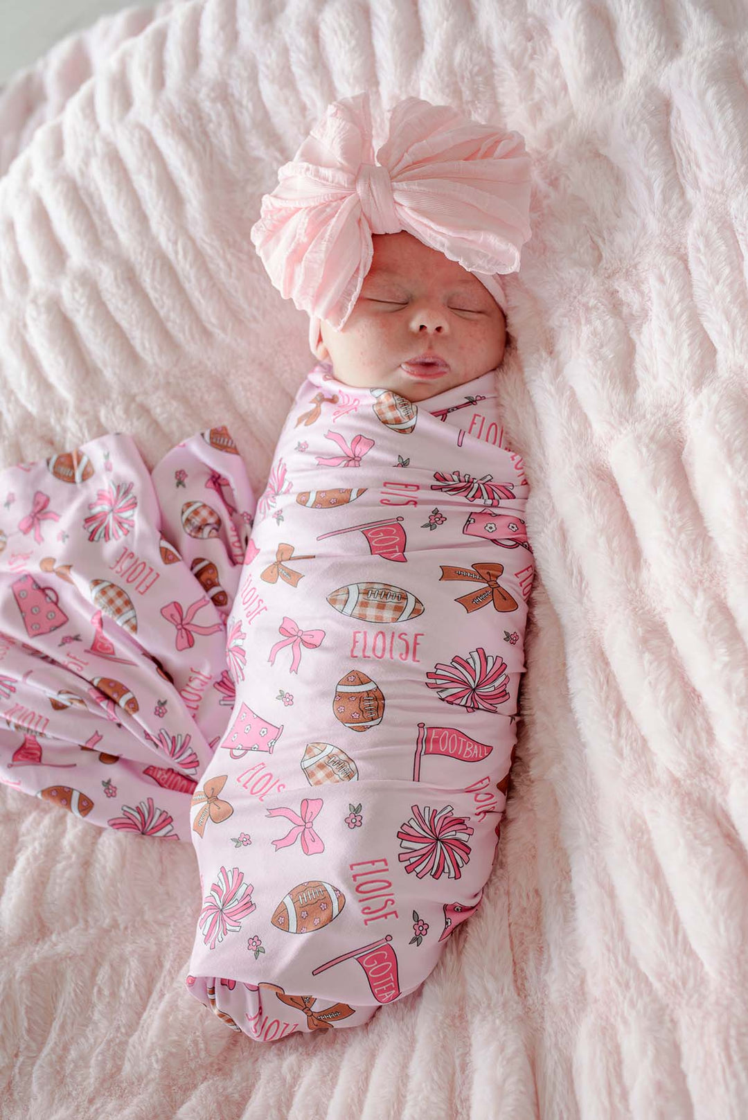 Sleeping newborn wearing a large pink bow headband, wrapped in a personalized Caden Lane 'Huddles and Cuddles' pink football and cheerleading print swaddle blanket with the name Eloise, lying on a pink furry blanket.