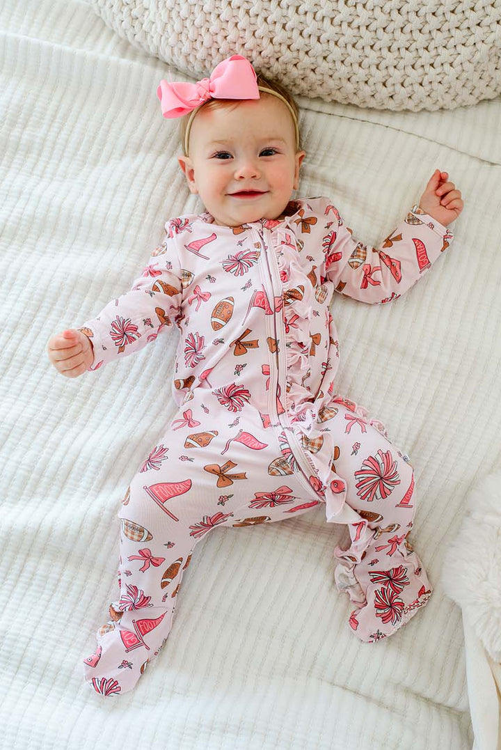 A baby lying on a white textured blanket, wearing a pink footed sleeper with a candy print and a matching pink bow headband.