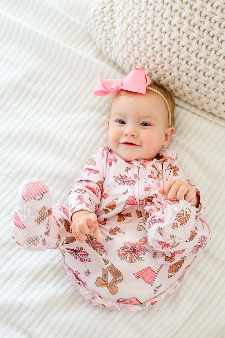 A baby lying on a white textured blanket, wearing a pink and white footed sleeper with a bow headband and matching booties.