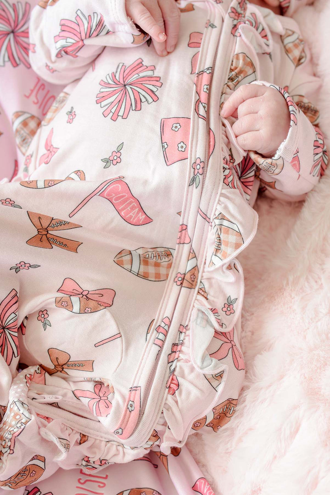A close-up of a baby wearing a pink and white zip-up sleeper with a high-heel and bow pattern, lying on a soft pink blanket.