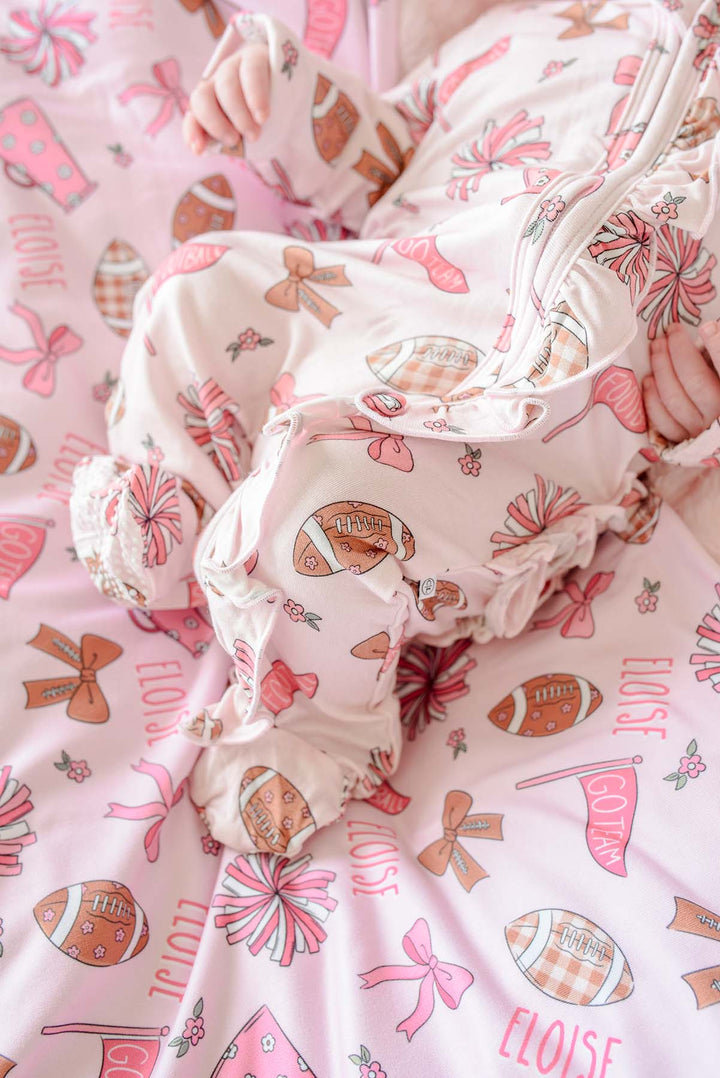 A close-up of a baby wearing a light pink footed sleeper with a brown and pink football and bow pattern, lying on a matching printed blanket.