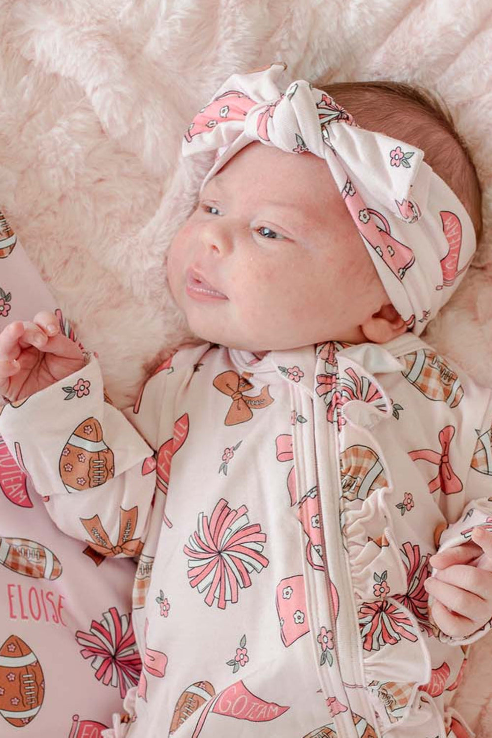 A baby lying on a fluffy cream blanket, wearing a cream zip-front footed sleeper with a matching headband and a pink patterned blanket underneath. The pattern features pink bows, brown footballs, and the word 'ELOISE'.