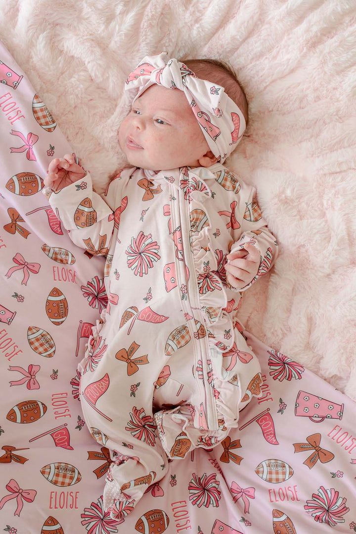 A baby lying on a fluffy cream blanket, wearing a cream zip-front footed sleeper with a matching headband and a pink patterned blanket underneath. The pattern features pink bows, brown footballs, and the word 'ELOISE'.