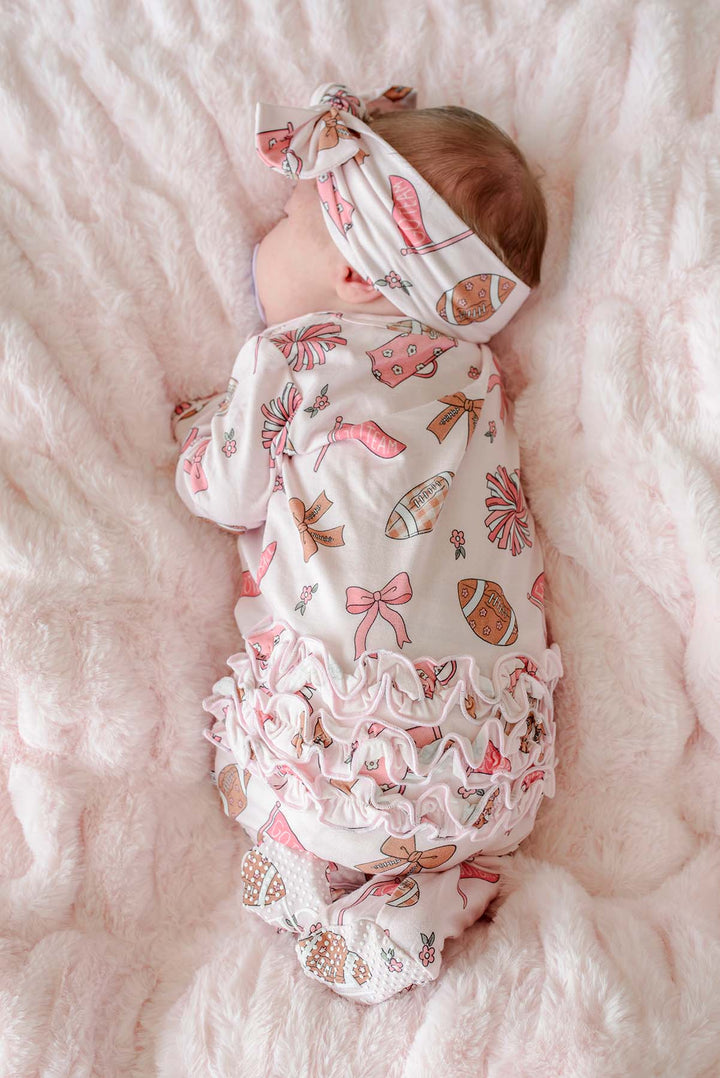 A baby sleeping on a fluffy pink blanket, wearing a white long-sleeve romper and matching headband with a pink and brown pattern of bows, hearts, and footballs.