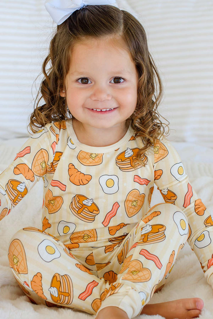 Toddler girl in Caden Lane I Love You A Brunch pajamas, seated, smiling, wearing a white bow