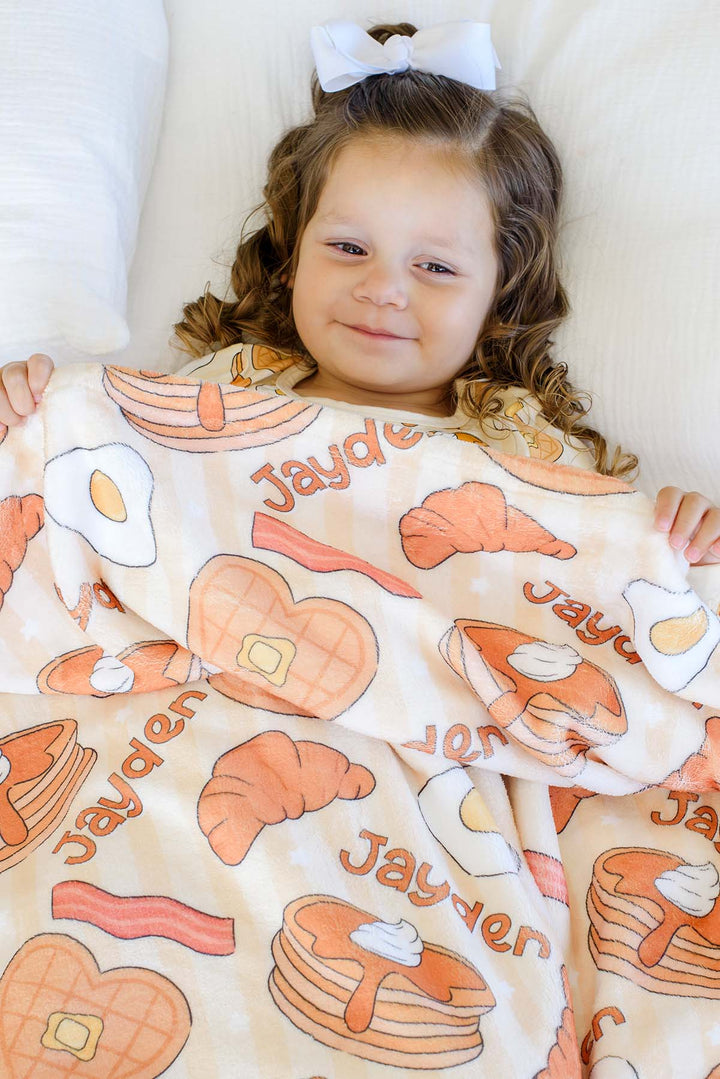 Toddler girl in bed holding up a Caden Lane personalized "Jayden" plush blanket, smiling