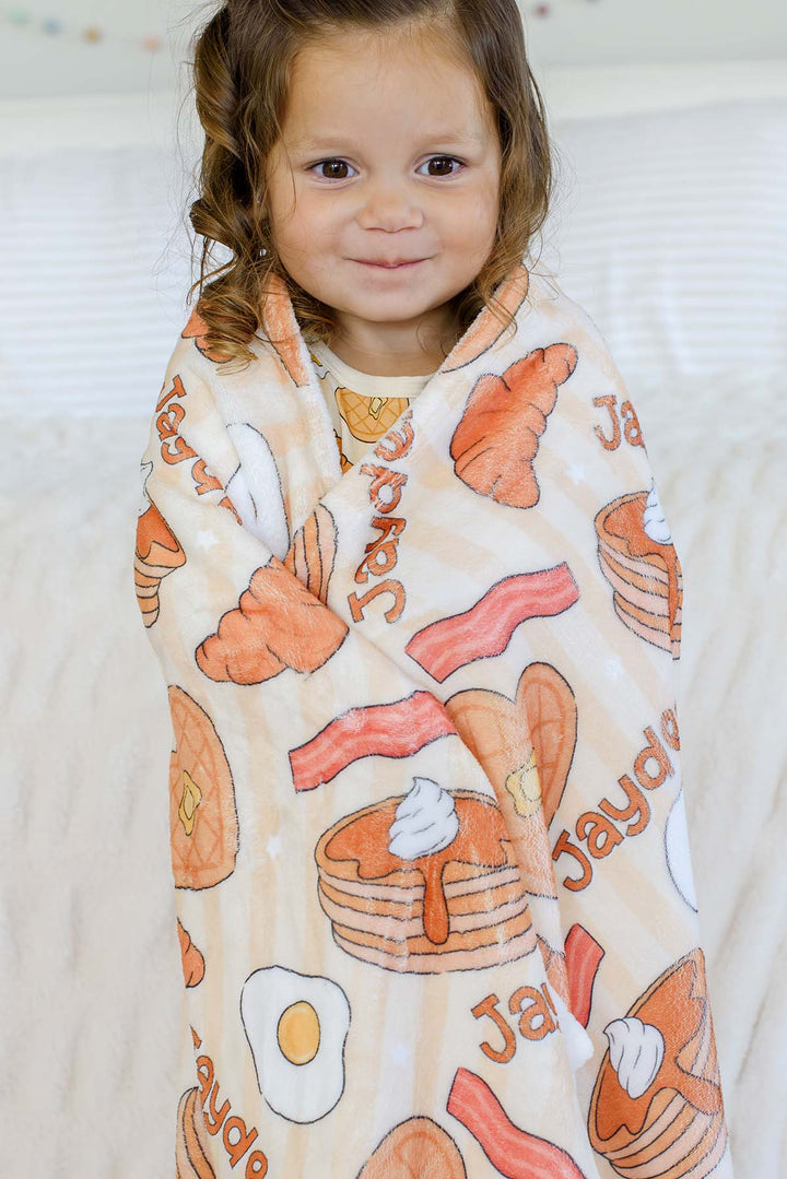 Toddler girl wrapped in a Caden Lane personalized "Jayden" plush blanket, smiling at camera