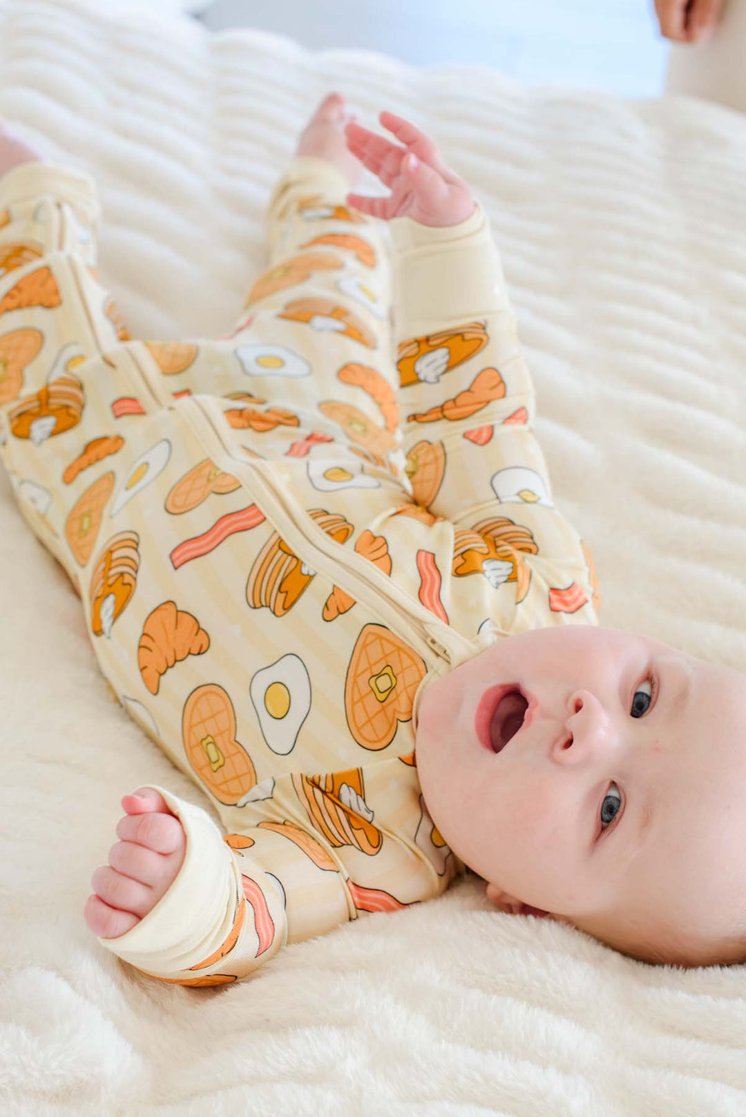 Baby lying on back in Caden Lane zip footie pajama with breakfast food print, pancake, egg, bacon, waffle and croissant print on cream stripes