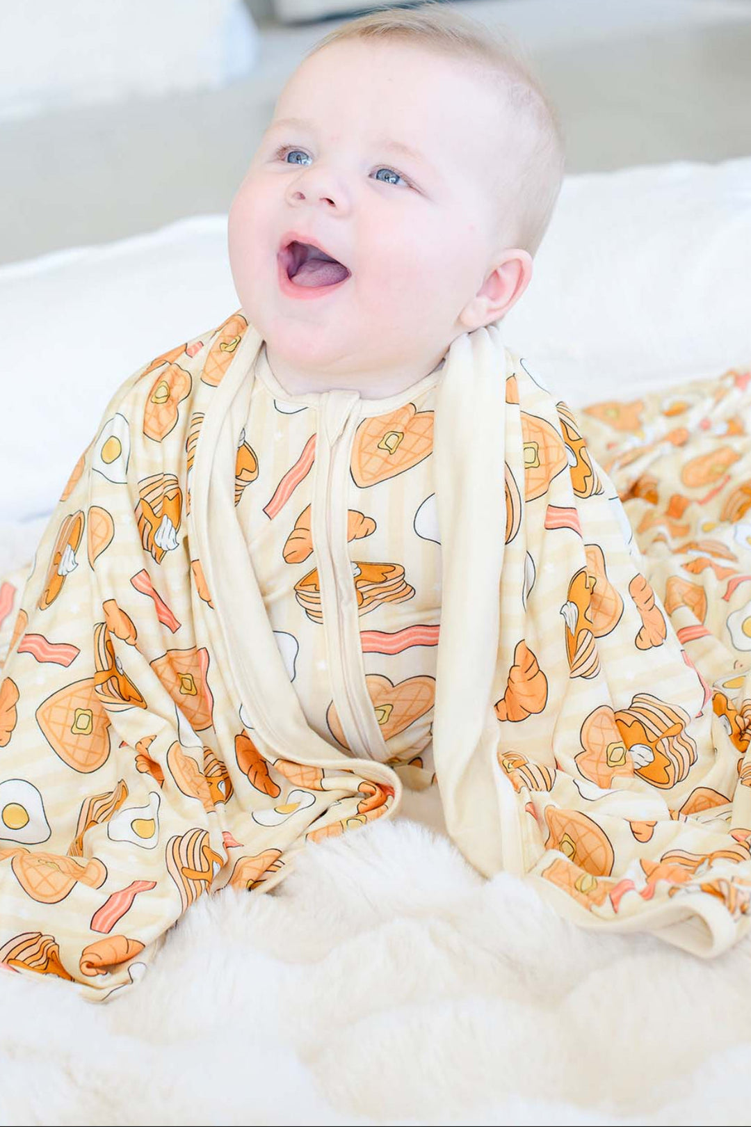 A baby wearing a Caden Lane ream-colored sleep sack with an orange and yellow breakfast pattern, sitting on a white fluffy rug in a bright room.