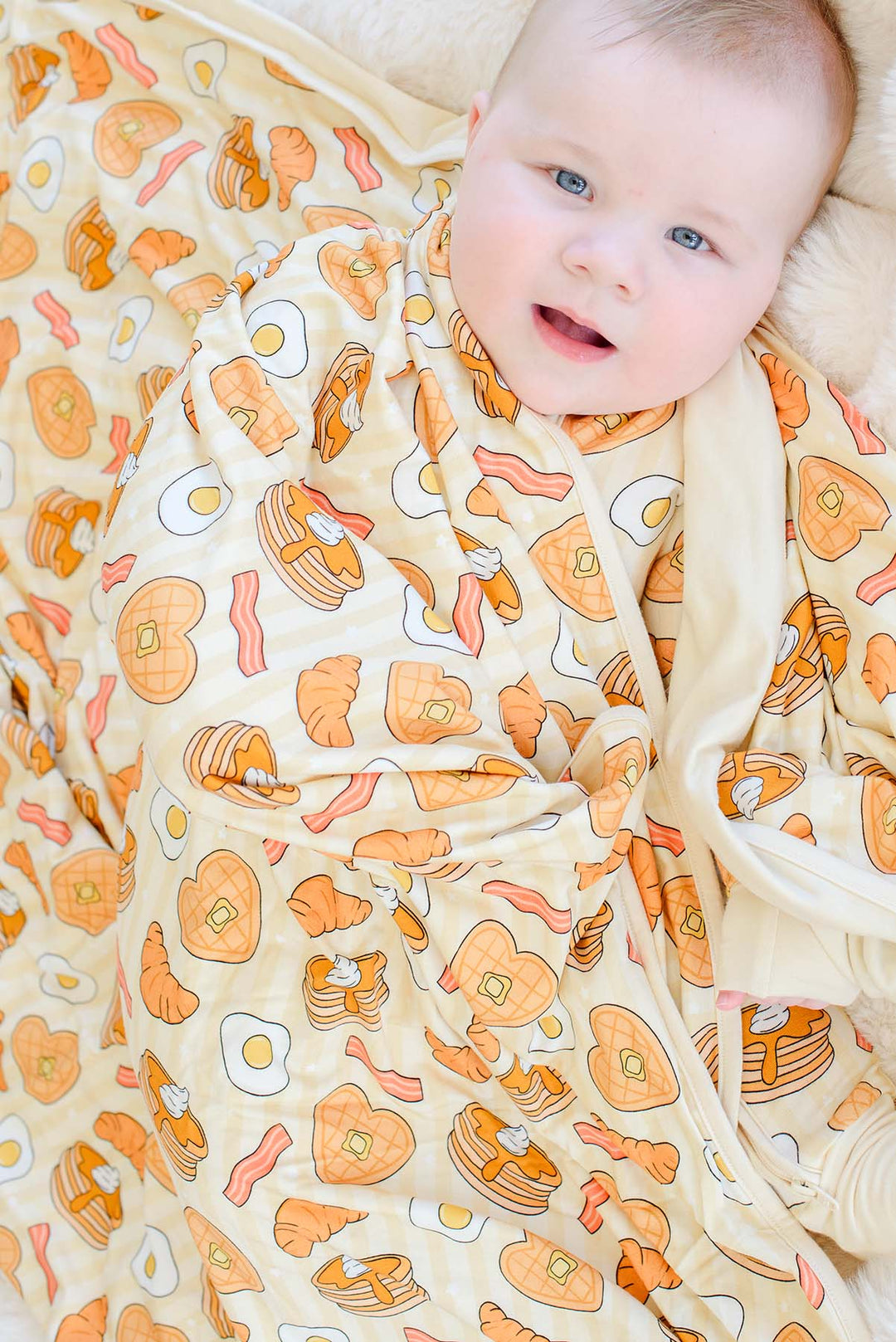 A baby wrapped in a cozy, cream-colored swaddle blanket with a breakfast-themed print of pancakes, bacon, eggs, and toast, lying on a soft beige blanket.