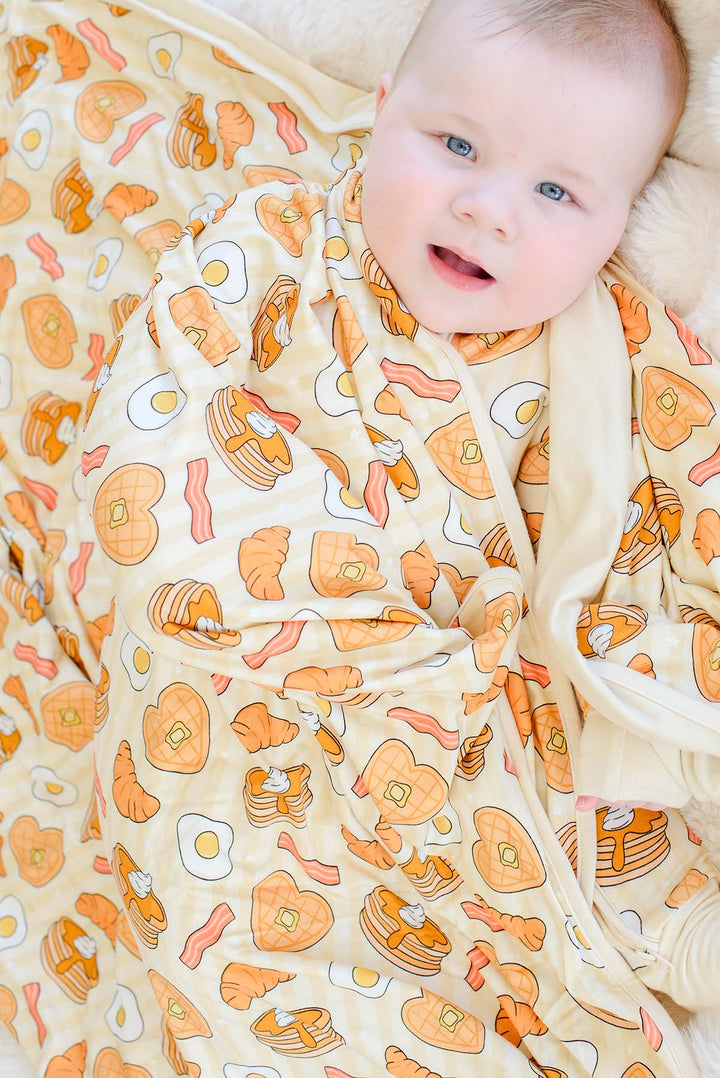A baby wrapped in a cozy, cream-colored swaddle blanket with a breakfast-themed print of pancakes, bacon, eggs, and toast, lying on a soft beige blanket.