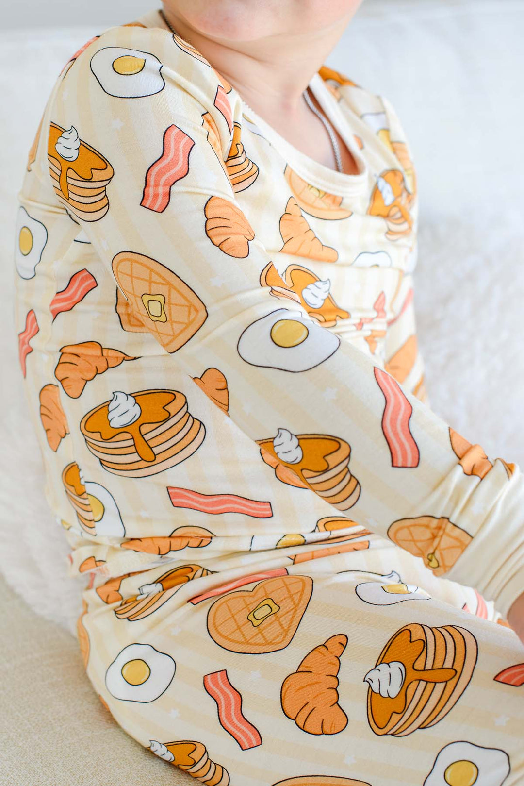 Close-up of child in Caden Lane I Love You A Brunch pajamas, showing pancake, egg, bacon and croissant print detail