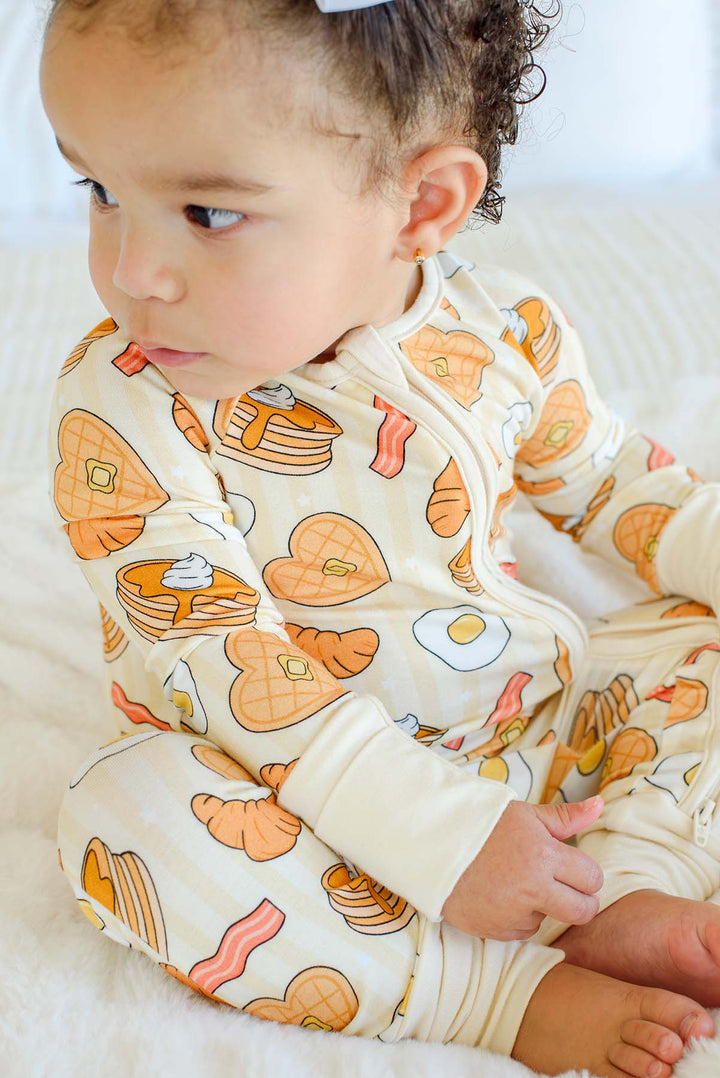 Close-up of toddler sitting in Caden Lane zip footie pajama, showing egg and waffle print