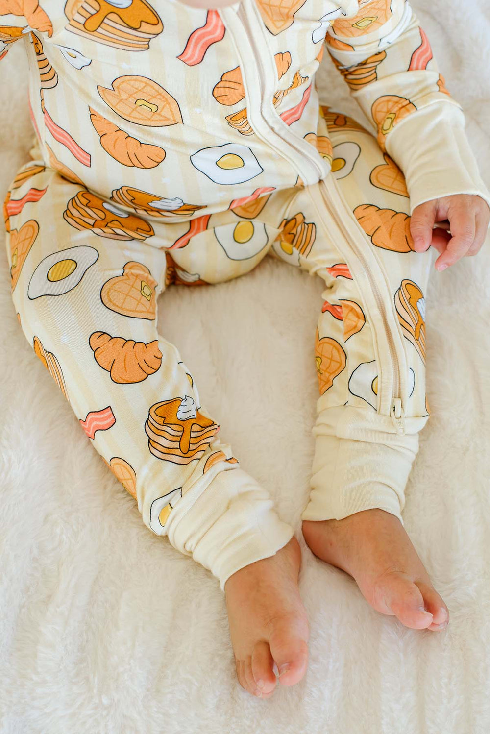 Close-up of toddler's legs and feet in Caden Lane zip footie pajama with breakfast food print, pancake, egg, bacon, waffle and croissant print on cream stripes