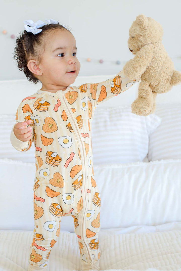 Toddler girl in white bow standing in Caden Lane zip footie pajama with pancake, egg and bacon print, holding a teddy bear